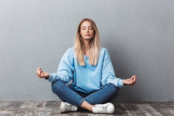 Pretty young blonde girl meditating while sitting in a yoga position with eyes closed isolated over gray background - Photo, Image