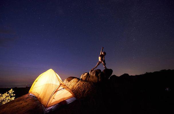 SVR camping in de bergen. Heldere toeristische tent op steile rots en jonge slanke vrouw poseren met opgeheven arm op donker blauwe sterrenhemel avondlucht. Toerisme, yoga, actieve levensstijl, klimmen concept. - Foto, afbeelding