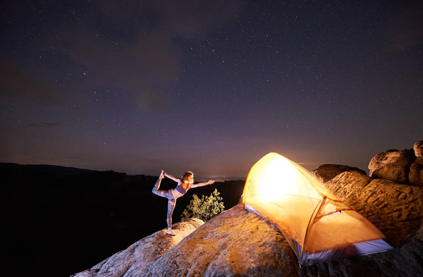 Frau in komplizierter Yoga-Pose auf einem Bein stehend auf steilen Felsen vor Touristenzelt am sternenklaren Abendhimmel und schwarzem Berghintergrund. Tourismus, aktiver Lebensstil, Campingkonzept. Narajasana - Foto, Bild