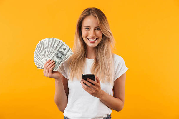 Retrato de uma jovem loira feliz mostrando um monte de notas de dinheiro e segurando o telefone celular isolado sobre fundo amarelo
 - Foto, Imagem
