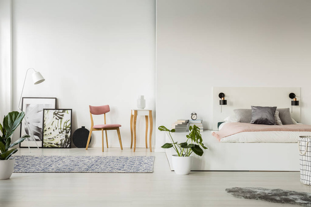 Real photo of a platform bed standing next to a chair, small table, posters and a lamp in spacious bedroom interior - Valokuva, kuva
