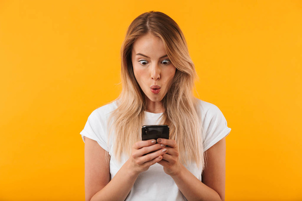 Portrait of a shocked young blonde girl looking at mobile phone isolated over yellow background - Foto, Imagen