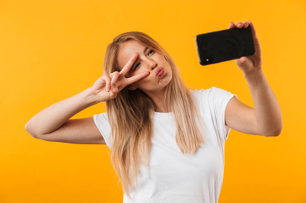 Happy young blonde girl showing peace gesture while taking a selfie with mobile phone isolated over yellow background - 写真・画像