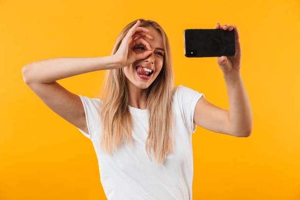 Happy young blonde girl showing ok gesture while taking a selfie with mobile phone isolated over yellow background - Фото, зображення