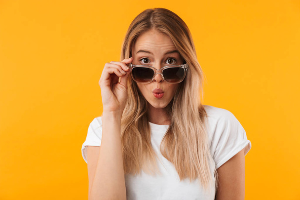 Portrait of a lovely young blonde girl in sunglasses posing while looking at camera isolated over yellow background - Foto, Bild