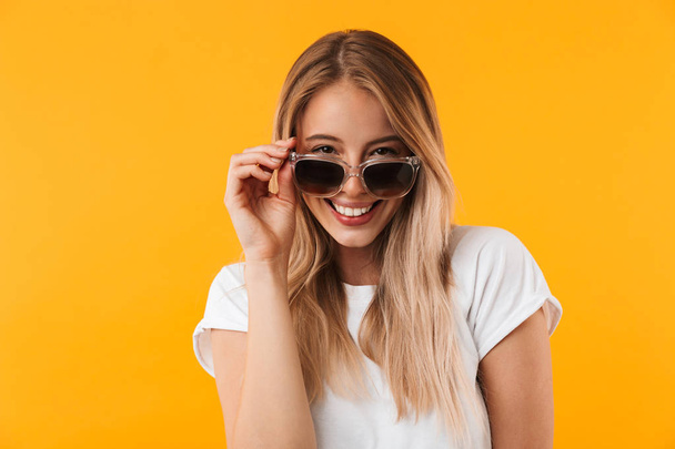 Portrait of a smiling young blonde girl in sunglasses posing while looking at camera isolated over yellow background - Fotó, kép