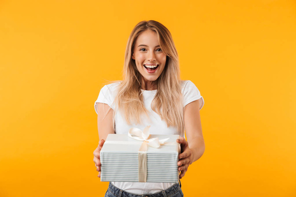 Portrait of a cheerful young blonde girl giving present box isolated over yellow background - Zdjęcie, obraz