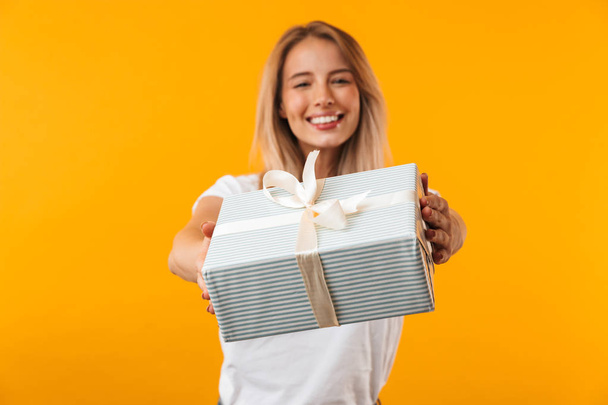 Portrait of a happy young blonde girl giving present box isolated over yellow background - Foto, Imagen
