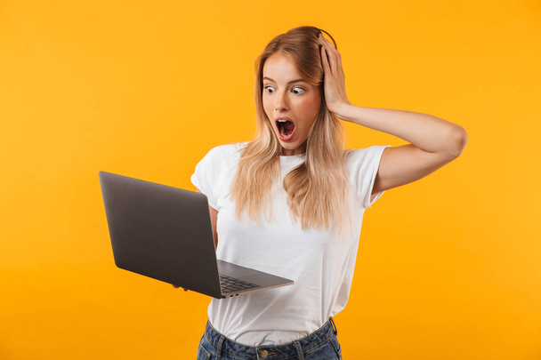 Portrait of a shocked young blonde girl holding laptop computer isolated over yellow background - Foto, Bild