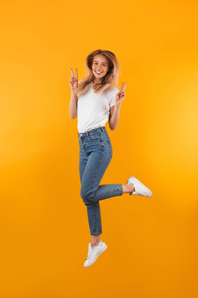 Full length portrait of a cheerful young blonde girl showing peace gesture while jumping isolated over yellow background - Foto, Imagem