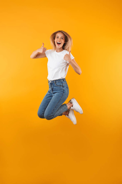 Full length portrait of a happy young blonde girl showing thumbs up gesture while jumping isolated over yellow background - Photo, image