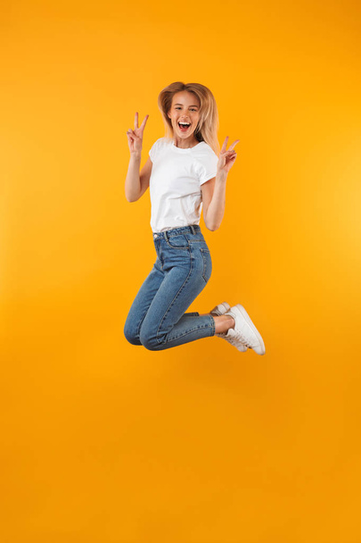 Full length portrait of a cheerful young blonde girl showing peace gesture while jumping isolated over yellow background - Foto, Imagen