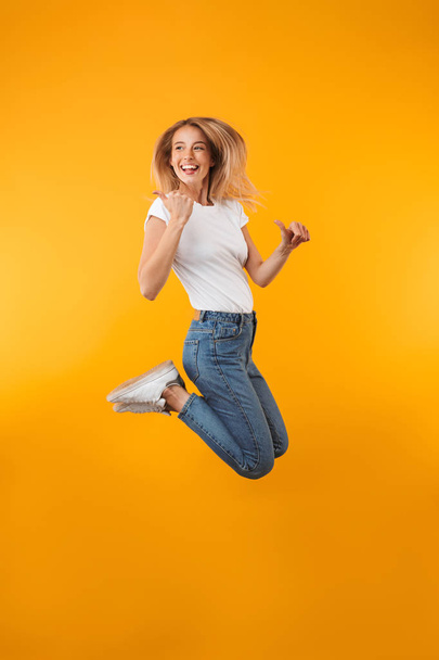Full length portrait of a cheerful young blonde girl pointing away while jumping isolated over yellow background - Фото, изображение