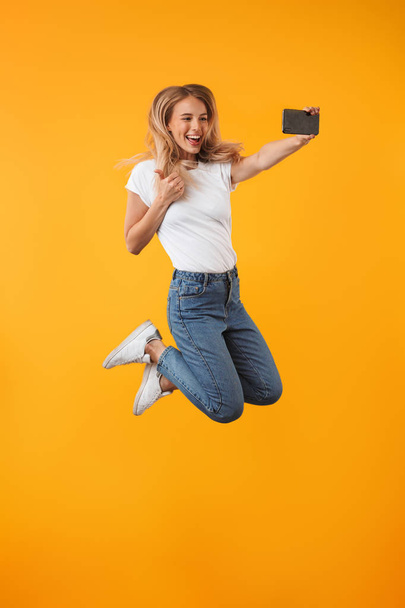 Full length portrait of a smiling young blonde girl showing thumbs up gesture while jumping and taking a selfie isolated over yellow background - Fotó, kép