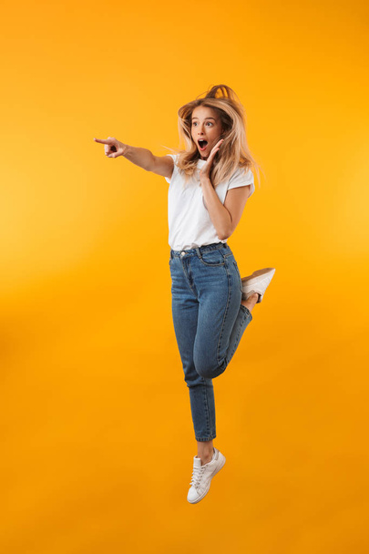 Full length portrait of a surprised young blonde girl pointing away while jumping isolated over yellow background - Fotó, kép