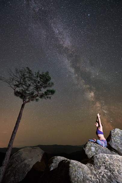 Όμορφη καλοκαιρινή νύχτα στα βουνά. Γυναίκα που κάθεται μόνη πάνω από τεράστιο ογκόλιθο που κάνει γιόγκα κοντά στο πεύκο, κάτω από καθαρό ουρανό με αναβοσβήνει αστέρια και Milky way. Εκδρομές και πεζοπορία έννοια. Σιντασάνα - Φωτογραφία, εικόνα