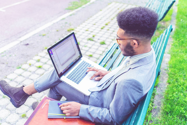 молодой современный черный бизнесмен сидит на скамейке под открытым небом с помощью ноутбука - портативный офис, удаленная работа, бизнес-концепция
 - Фото, изображение