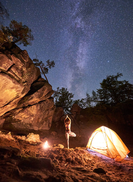 Noche acampando entre montañas rocas. Lit by campfire back view silhouette of slim woman standing on one leg with raised arms doing yoga at tourist tent on clear starry sky background. Vrikshasana
 - Foto, imagen