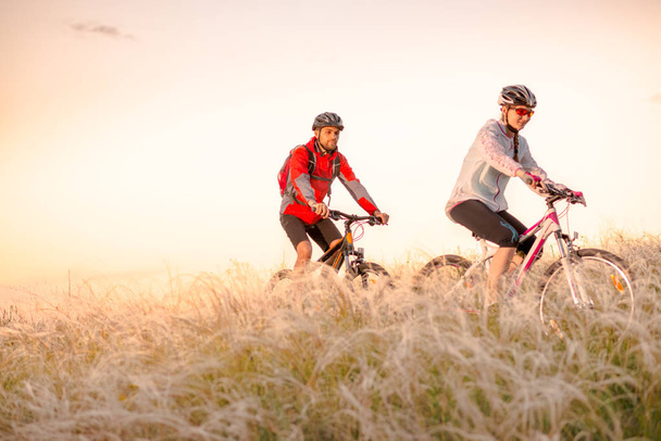 Young Couple Riding Mountain Bikes in the Beautiful Field of Feather Grass at Sunset. Adventure and Family Travel. - Photo, image