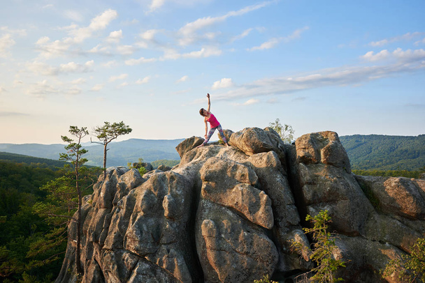 Vonzó sportos lány csinál bonyolult jóga gyakorlatok, tetején hatalmas rakás sziklák a zöld hegyek és a tiszta kék ég háttér. Hegymászás, a turizmus, a fitness és az egészséges életmód fogalma. - Fotó, kép