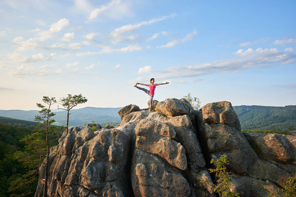 Attrayant mince fille touristique faisant des exercices de yoga sur le dessus de roche énorme sur les montagnes vertes et fond ciel bleu clair le matin d'été. Sport, yoga, fitness et mode de vie sain concept
. - Photo, image