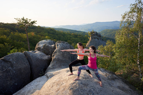 Two slim athletic barefooted girls doing yoga exercises on top of huge rocks on green mountains and clear blue sky background. Climbing, tourism, fitness and healthy lifestyle concept. Virabhadrasana - Photo, Image