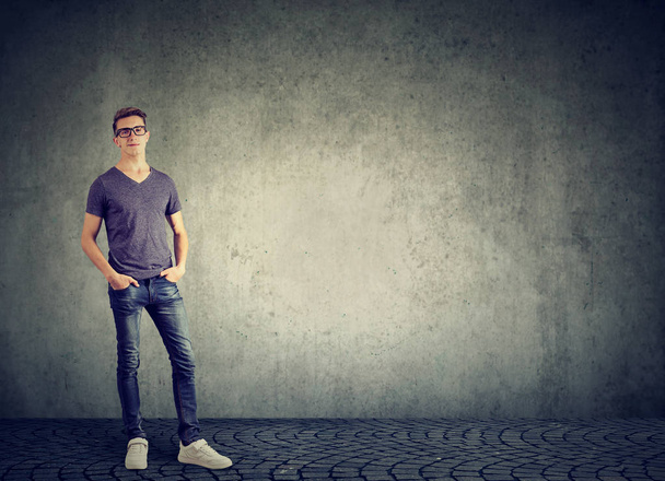 Young casual man wearing jeans with t-shirt and glasses standing with hands in pockets against gray wall - Fotoğraf, Görsel