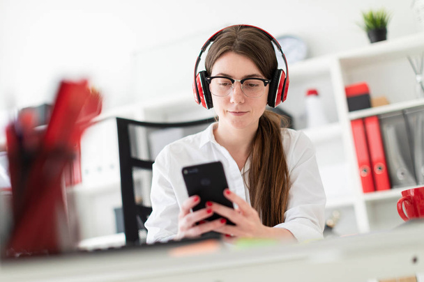 A young girl sits in headphones at a table in the office and holds a phone in her hand. - Foto, imagen