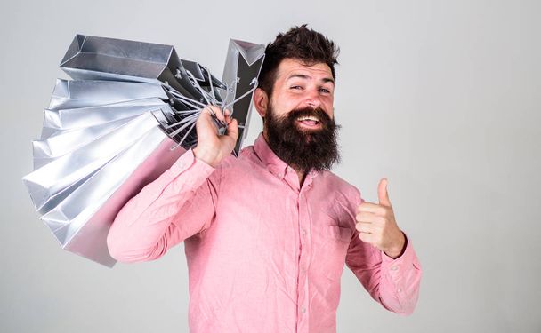 Black friday concept. Guy shopping and shows thumbs up gesture. Hipster on happy face recommends to buy. Man with beard and mustache carries bunch of shopping bags, grey background - Photo, Image