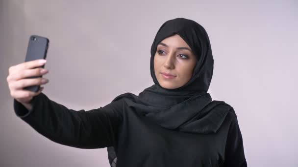 Young weet muslim girl in hijab is making selfie on her smartphone, watch photos, communication concept, religious concept, grey background. - Video