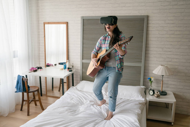 Asian female musician playing wooden guitar on her bed and wearing the VR. She used her VR device to Share her skill with her fans on social website. - Фото, зображення