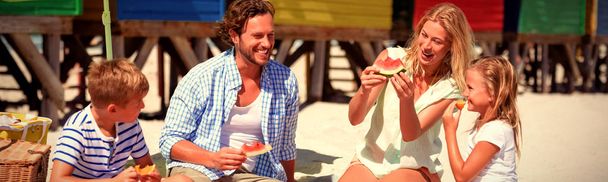 Happy family eating watermelon while sitting together on blanket at beach during sunny day - Valokuva, kuva