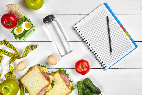 Το Top view σχεδιασμός notebook με αντίγραφο χώρου και υγιεινό πρωινό. Σάντουιτς με ζαμπόν και τυρί, αυγά, αγγούρι, ντομάτα και μπουκάλι νερό στο λευκό ξύλινο τραπέζι. - Φωτογραφία, εικόνα
