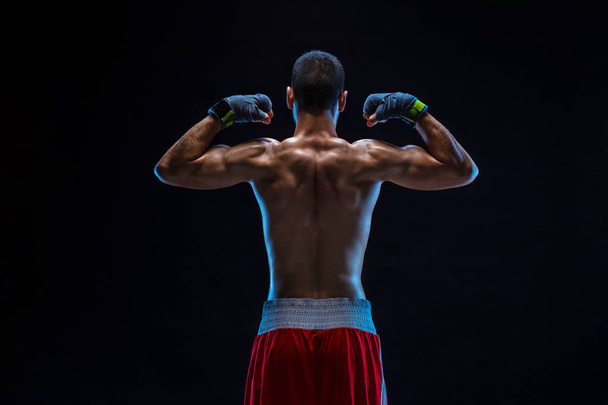 A boxer in his shorts stands with his back on black background. Handsome young bodybuilder with toned body posing shirtless - Foto, Imagen