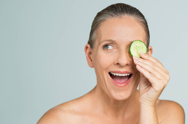 Portrait of mature beautiful naked woman smiling hiding eye behind cucumber slice. Beauty portrait of a smiling senior woman isolated on gray background. Funny senior woman in a playful mood covering eyes with cucumber slice. - Valokuva, kuva
