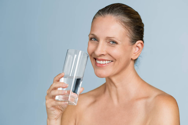 Portrait of happy mature woman holding a glass of filtered water isolated on grey background. Cheerful senior woman holding half filled glass of water and looking at camera. Health care concept and hydration for perfect beauty skin. - Fotó, kép