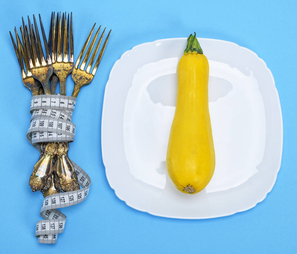 yellow squash on a white ceramic plate, next to a pile of iron forks wrapped with measuring tape, blue background - Photo, Image