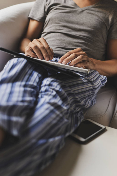 closeup of a young caucasian man in pajamas sitting in a comfortable armchair using a keyboard and a digital tablet - Fotó, kép
