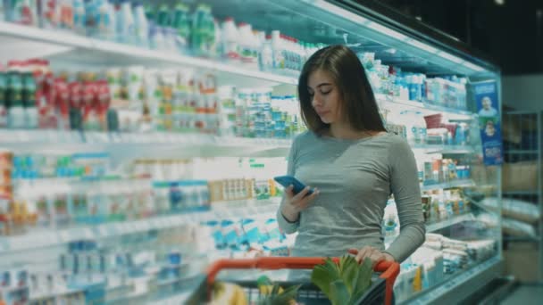 Χαρούμενα νεαρή όμορφη γυναίκα που μιλάει στο τηλέφωνο χαμογελώντας ενώ αγοράζουν τρόφιμα στο σούπερ μάρκετ copyspace κατάστημα παντοπωλείο ψώνια αγοραστή καταναλωτισμός πελάτης τεχνολογία επικοινωνίας καλώντας - Πλάνα, βίντεο