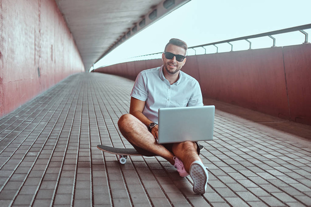 Πορτρέτο του happy όμορφος μοντέρνος επαγγελματίας σε γυαλιά ηλίου ντυμένος με ένα άσπρο πουκάμισο και σορτς που εργάζονται σε ένα φορητό υπολογιστή, ενώ κάθεται σε ένα skateboard κάτω από τη γέφυρα, βλέπουν μια φωτογραφική μηχανή. - Φωτογραφία, εικόνα