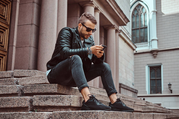 黒のジャケットとジーンズのヨーロッパの古い建物に対しての手順の上に座ってスマート フォンを使って服を着たファッショナブルな男. - 写真・画像