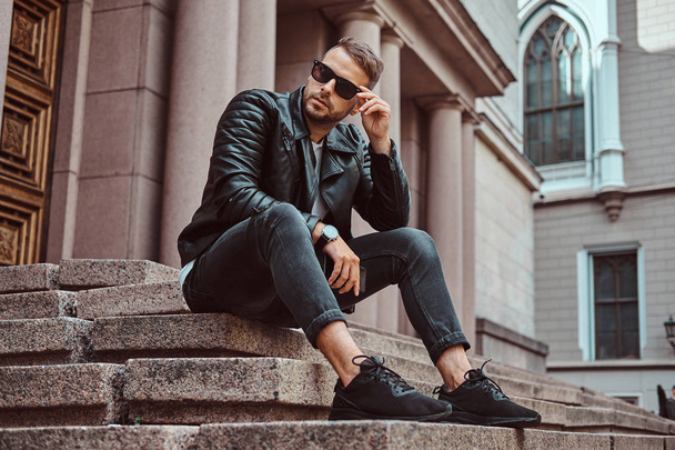 Modieuze man gekleed in een zwarte jas en jeans heeft een smartphone zit op stappen tegen een oud gebouw in Europa. - Foto, afbeelding