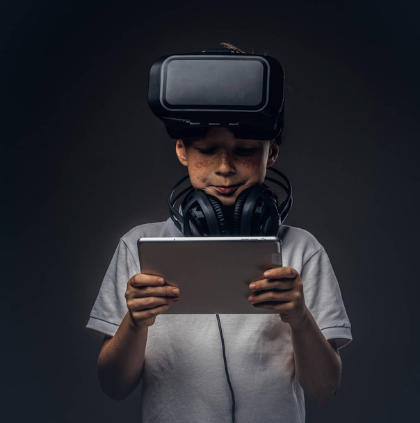 Portrait of a little boy dressed in a white t-shirt, with virtual reality glasses and headphones and using a digital tablet at a studio, isolated on dark background. Education and technology concept. - Photo, Image