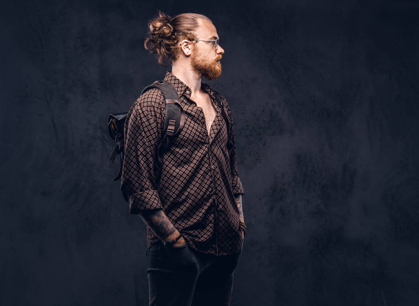 Portrait of a redhead hipster student in glasses dressed in a brown shirt, holds a backpack, posing at a studio. Isolated on a dark background.  - Foto, imagen