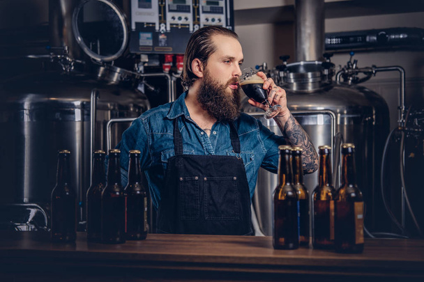 Bebaarde getatoeëerd hipster man in een spijkerbroek shirt en schort werken in een fabriek van de brouwerij, die achter een teller, drinkt een ambachtelijke bier voor de kwaliteitscontrole. - Foto, afbeelding