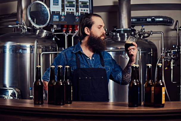 Bearded tattooed hipster male in a jeans shirt and apron working in a brewery factory, standing behind a counter, drinks a craft beer for quality control. - Photo, Image