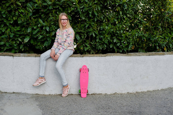  muotokuva vaalea tyttö vaaleanpunainen lippis ja rullalauta puistossa. Teini-ikäinen tyttö silmälaseissa istuu ulkona kadulla. Lapsuus, kesä, loma, urheilu
  - Valokuva, kuva