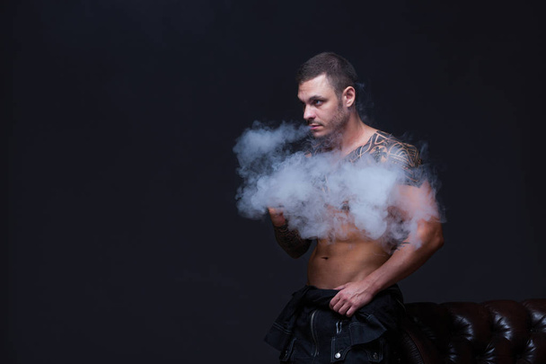 Vaper. The man with a muscular naked torso with tattoos smoke an electronic cigarette on the dark background - Фото, изображение
