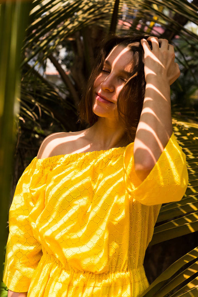 Beautiful young girl in yellow dress posing under palm leaves - Фото, зображення