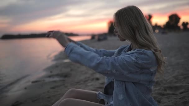 Mujer joven sentada en la playa del río al atardecer mirando alrededor y soñando
 - Metraje, vídeo